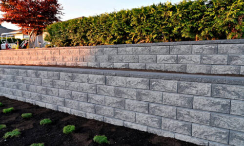 A close-up capture of a gray colored block stone retaining wall built as a two tier wall into an existing garden landscape viewed from the side in the early morning hours under sunny conditions.