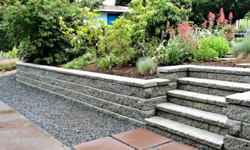 A retaining wall with built in steps into an existing garden landscape in gray color with brown stepping stones leading up to the staircase.