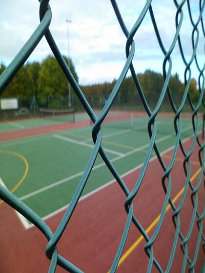 Green hard tennis court viewed through a chain link fence.