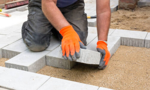 Hands of a builder laying new paving stones carefully placing one in position on a levelled and raked soil base.